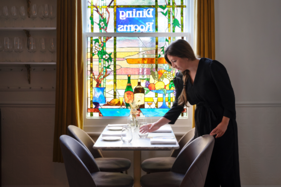 Server laying a table beside a window and a shelf of colourful bottles, Cambridge